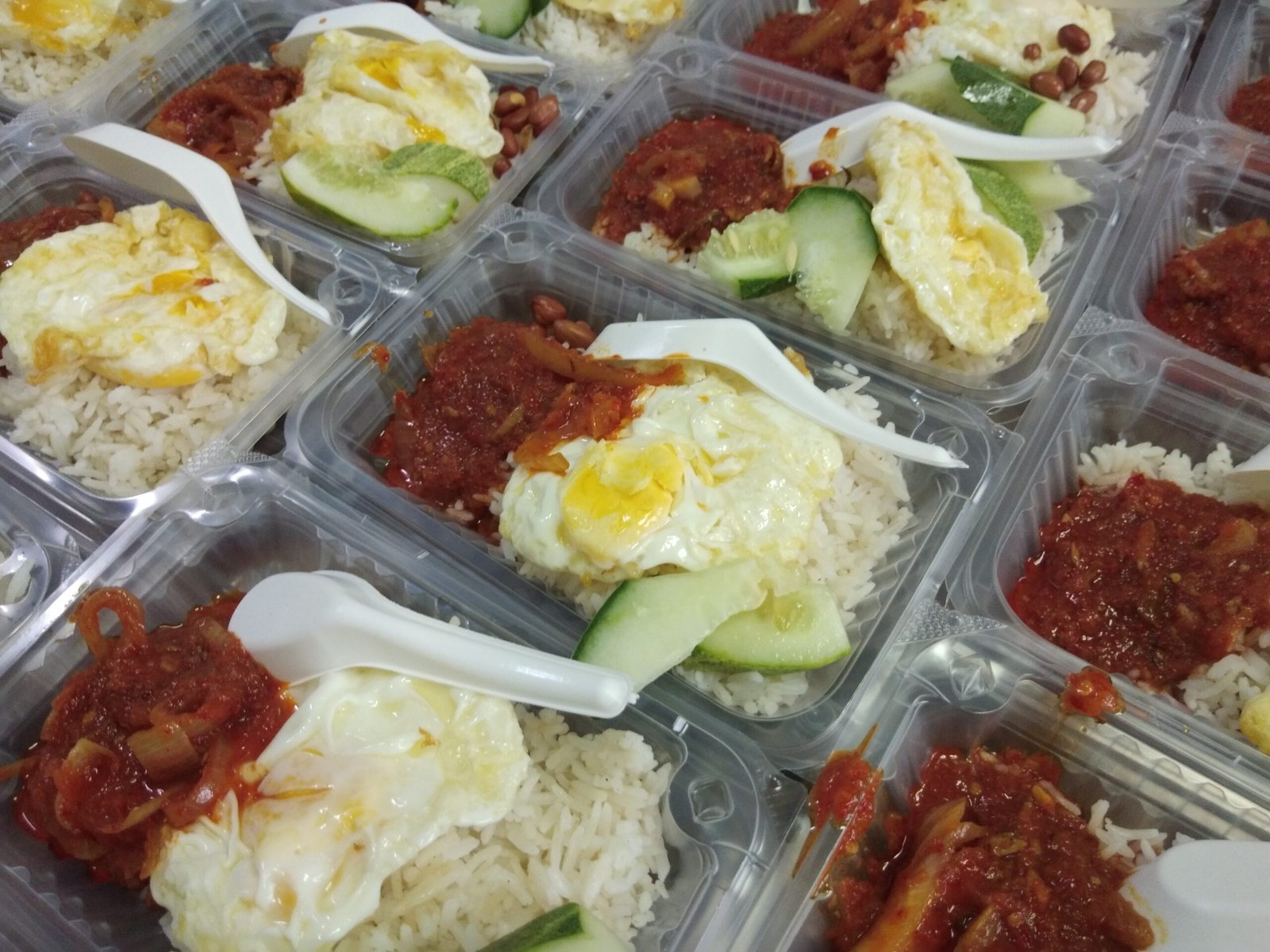Nasi lemak di dalam bekas dengan telur goreng, timun dan sambal.