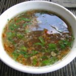 Semangkuk sup ayam jernih bertabur daun hijau dan bawang goreng.